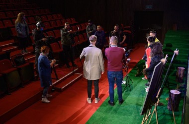 przed sceną konferencja prasowa po prawej na scenie siedzi czwórka aktorów po prawej siedmiu dziennikarzy z mikrofonami i dwie dziennikarki