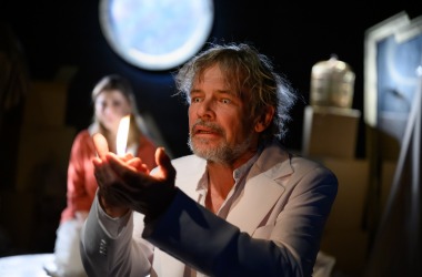 Aktor (Anioł) w białym garniturze trzyma świeczkę w dłoni. W tle aktorka patrzy na niego