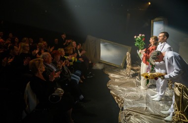 Po lewej stronie publiczność bije brawo, po prawej aktorzy kłaniają się widzom, trzymają kwiaty