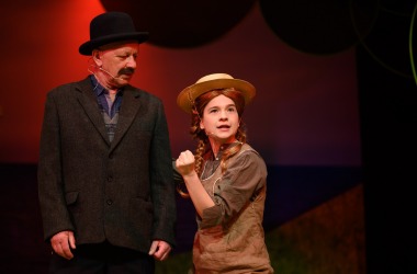 po prawej Ania młoda dziewczyna w rudej peruce kapeluszu ma zawzięta minę, zaciśnięta pięść, obok niej po lewej Mateusz starszy mężczyzna w ciemnym garniturze czarnych wąsach i czarnym kapeluszu