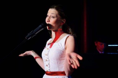 młoda aktorka w białej sukience w jasnych włosach śpiewa do mikrofonu z tyłu za nią półmroku siedzi pianista