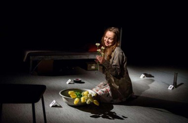 Zdjęcie aktorki, siedzi na scenie, w ręce trzyma rumianek, na podłodze przed nią są żółte tulipany