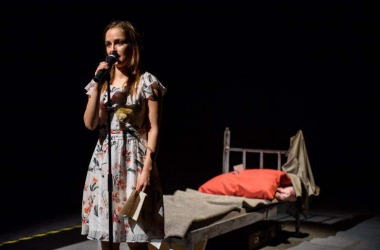 Zdjęcie aktorki, stoi przed mikrofonem, za nią stoi łóżko