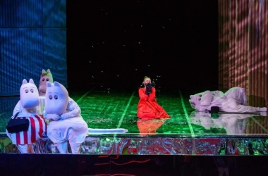 Na środku sceny klęczy Mała Mi (aktorka ubrana w czerwony kostium), na rękach ma czarne rękawiczki przykłada do twarzy. Po lewej stronie siedzą Mama Muminka, Tata Muminka, Migotka, po prawej stronie Muminek