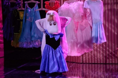 Aktorka w białym kostiumie (Migotka) przymierza niebieską sukienkę. w tle za nią wisi na wieszakach kilka kolorowych sukienek