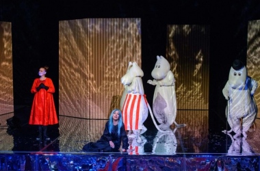 Po lewej stronie Mała Mi w czerwonym kostiumie, na środku siedzi Bufka w czarnym kostiumie i niebieskiej peruce. Z tyłu za nią stoją Mama Muminka, Muminek i Tata Muminek