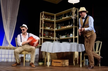scena w kuchni, po prawej mężczyzna w słomkowym kapeluszu gra na gitarze i tańczy, po lewej stronie na krześle siedzi młody mężczyzna udaje, ze gra na wiadrze, ma na twarzy okulary słoneczne