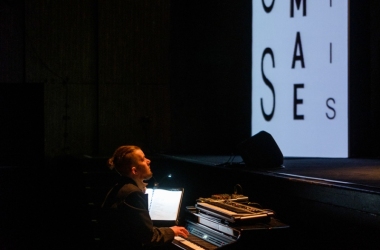 na środku przed sceną siedzi przed pianinem młody mężczyzna ubrany  w czarną marynarkę , na scenie litery o różnych wielkościach na białym tle czarna czcionka