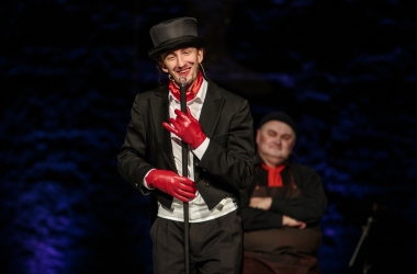 na środku młody aktor ubrany w czarny frak i czarny melonik trzyma  czarną laskę na dłoniach ma czerwone rękawiczki