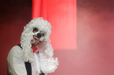 na scenie czerwone światło dym sceniczny po lewej siedzi młody aktor w białej koszuli i czarnej kamizelce na głowie ma długą białą perukę i maskę biały pysk psa patrzy w stronę widzów