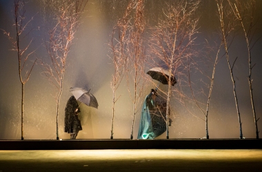 wiatr porusza drzewami, jest burza, między drzewami dwie postacie w pelerynach i parasolkami próbują iść
