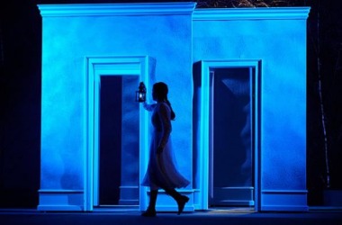 niebieskie światło, dekoracja imitująca ściany, na środku młoda kobieta trzyma w ręce lampę naftową przechodzi między ścianami