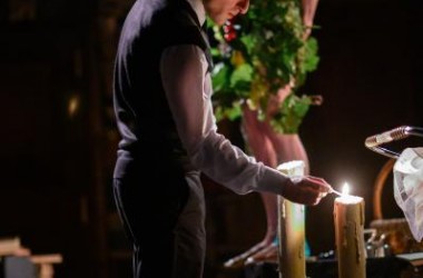Na pierwszym planie mężczyzna zapala dwie świeczki, w tyłu za nim mężczyzna nagi, ma na biodrach kwiaty