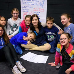 na scenie ośmioro dzieci siedzi na podłodze na środku pomiędzy nimi aktorka z tyłu za nimi duża kartka z zapiskami