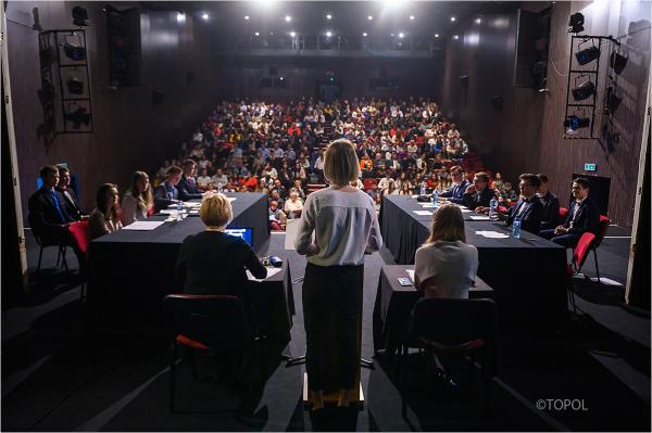 duża scena teatru widok siedzących widzów na drugim planie na pierwszym planie kobieta widok od tyłu blondynka w białej koszuli czarnej spódnicy po lewej i prawej stronie stoliki przy których siedzi kilkanaście młodych ludzi