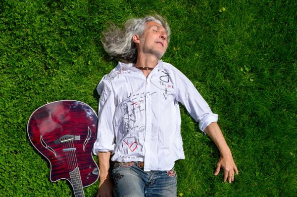 na zielonej trawie leży zadowolony wojtek klich muzyk z gitara ma długie siwe włosy i biała koszulę