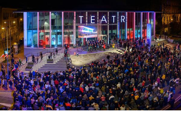 przed schodami teatru stoi kilkaset osób maja śpiewniki w ręce na schodach stoi kilkunastu chórzystów