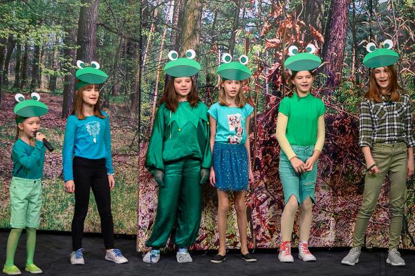 sześć dziewczynek wieku 10 lat ubrane na żółto jak żabki śpiewają w tle dekoracja ścianka imitująca las