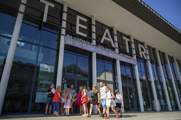 Przed wejściem do teatru stoi kilkanaście dzieci z rodzicami, u góry duży napis Teatr