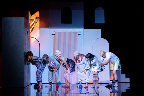 w rzędzie ustawieni pochyleni sześciu aktorów trzy kobiety i trzech mężczyzn ubrani w kolorowe piżamy stoją przy drzwiach i podsłuchują u góry nad nimi oświetlony na żółto aktor z wąsami patrzy na nich