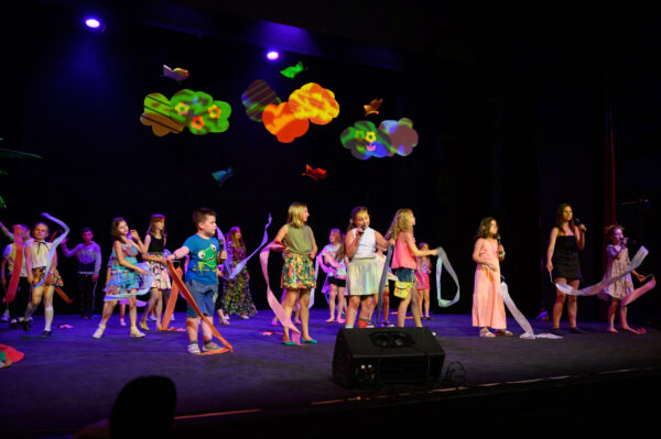 na scenie piętnaścioro dzieci ubrani w kolorowe ubrania, uśmiechają się, tańczą