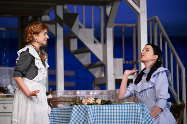 na pierwszym planie dwie aktorki. Po lewej aktorka gra Anię Shirley ma rudą perukę i jasno niebieską sukienkę. Na środku stolik po prawej stronie siedzi aktorka grająca Dianę przyjaciółkę Ani. Uśmiechają się do siebie. Z tyłu fragment schodów