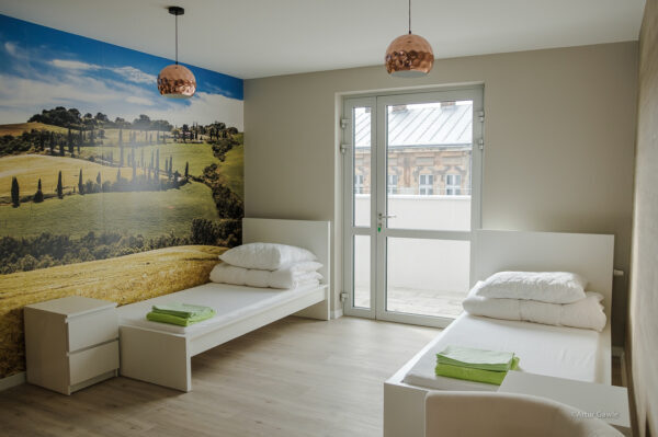 pokój po lewej i prawej białe łóżka między nimi drzwi balkonowe po lewej na ścianie fototapeta pagórek zielony, drzewa