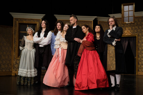 Czterech aktorów i cztery aktorki w kolorowych historycznych strojach i perukach stoją przed widzami