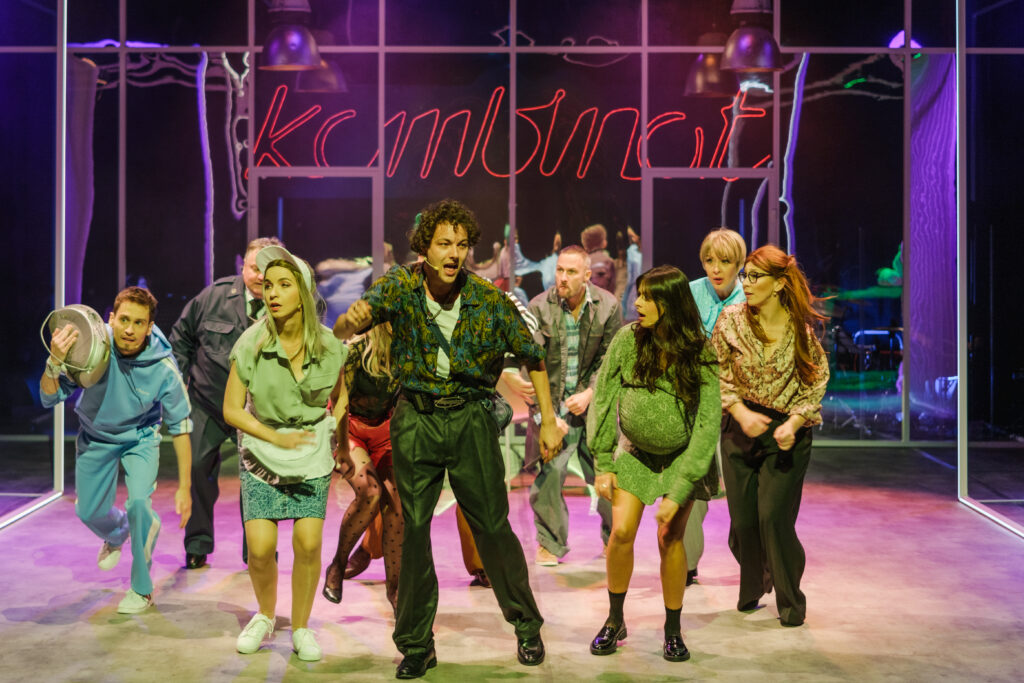 dziewięć aktorek i aktorów tańczy na tle napis kombinat